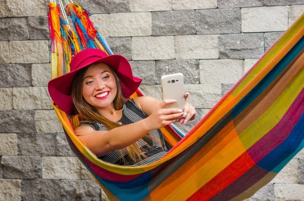 Primer plano de la joven hermosa mujer con un sombrero de moda rojo y relajarse en una hamaca y tomar una selfie con su teléfono celular, en un fondo borroso — Foto de Stock