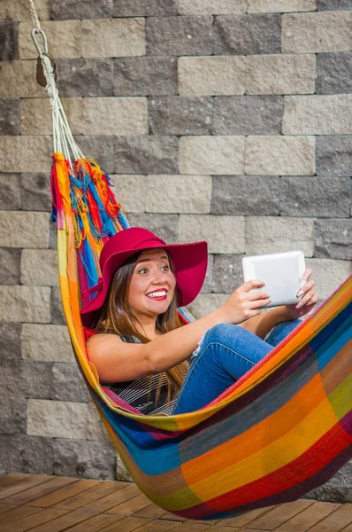 Close up van jonge mooie vrouw draagt een rode mode hoed en ontspannen in een hangmat en het nemen van een selfie met haar tablet, in onscherpe achtergrond — Stockfoto