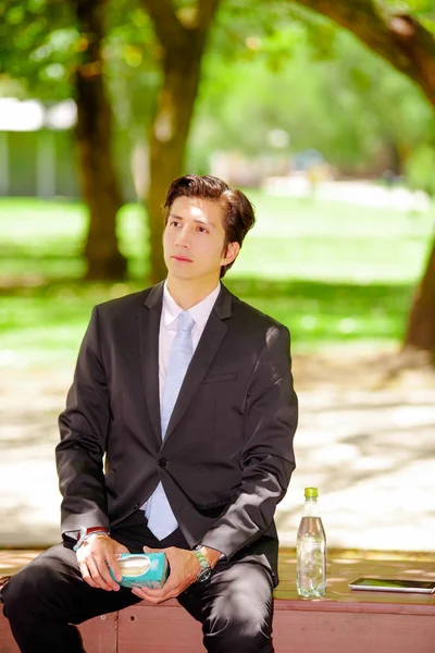 Young businessman wearing a suit and holding a sandwich inside of a board box at outdoors, in a blurred park background — Stock Photo, Image