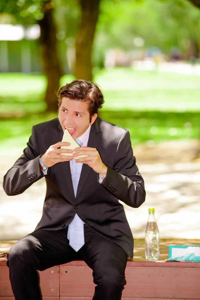 Bonito jovem empresário vestindo um terno e fazendo uma enorme mordida comendo um sanduíche ao ar livre, em um fundo de parque borrado — Fotografia de Stock