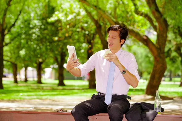 Gutaussehender junger Geschäftsmann im Anzug, der ein Sandwich isst und mit der anderen Hand ein Tablet im Freien hält, vor verschwommenem Parkhintergrund — Stockfoto
