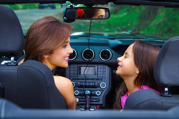 Närbild av vacker kvinna och hennes underbara dotter iklädd en rosa klänning som poserar inne i en svart lyxbil på en roadtrip, i en suddig natur bakgrund — Stockfoto