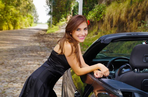 Gros plan d'une belle femme souriante portant une robe noire et une fleur rouge dans sa tête et posant devant une voiture noire de luxe lors d'un road trip, la voiture debout sur la ligne de touche, dans une nature floue — Photo