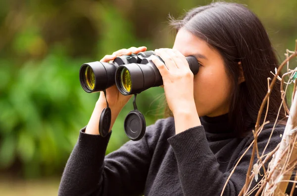 Young woman looking through black binoculars in the forest in a blurred background — Stock Photo, Image