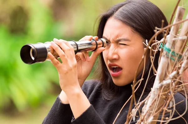 Surprised young woman looking through black monocular in the forest in a blurred background — Stock Photo, Image