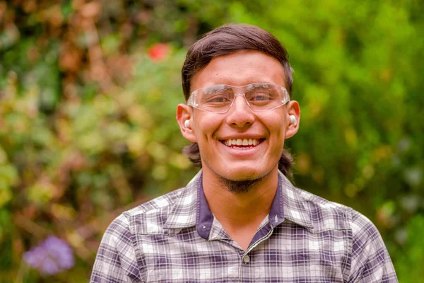 Retrato de jovem trabalhador sorrindo usando óculos de segurança transparentes, camisa de manga longa e tampões de ouvido, em um fundo de natureza turva — Fotografia de Stock