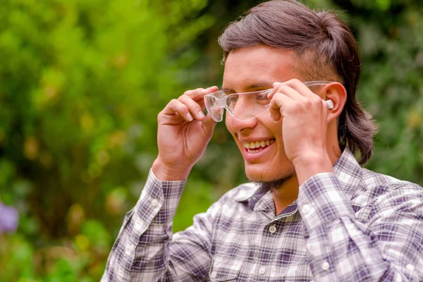 Retrato de jovem trabalhador vestindo óculos de segurança transparentes, camisa de manga comprida e tampões de ouvido para proteger do ruído, em um fundo de natureza turva — Fotografia de Stock