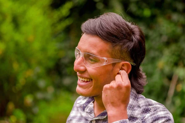 Retrato de jovem trabalhador sorrindo usando óculos de segurança transparentes, e vestindo uma camisa de manga longa, colocando tampões de ouvido para proteger do ruído, em um fundo de natureza turva — Fotografia de Stock