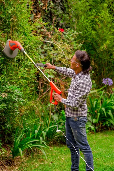 Close up of young worker wearing transparent glasses protection and holding a lawn trimmer mower cutting some bushes, in a blurred nature background — Stock Photo, Image