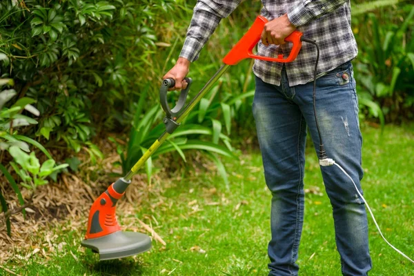 Unga arbetstagare klädd i jeans och långärmad tröja och med en gräsmatta trimmer klipparen klippa gräs i en suddig natur bakgrund — Stockfoto