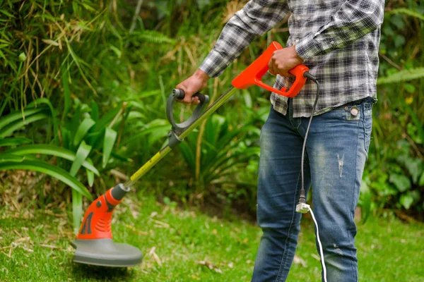 Trabajador joven que usa jeans y camisa de manga larga y usa una cortadora de césped cortando hierba en un fondo de naturaleza borrosa — Foto de Stock