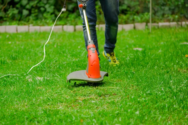 Young worker wearing jeans and using a lawn trimmer mower cutting grass in a blurred nature background — Stock Photo, Image