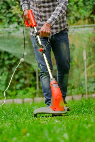 Молодой рабочий с газонокосилкой стрижет траву на размытом природном фоне — стоковое фото