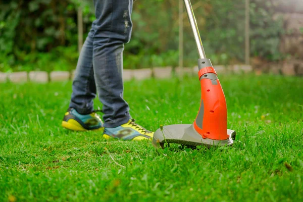 Närbild av unga arbetstagare bära jeans och med en gräsmatta trimmer klipparen klippa gräs i en suddig natur bakgrund — Stockfoto