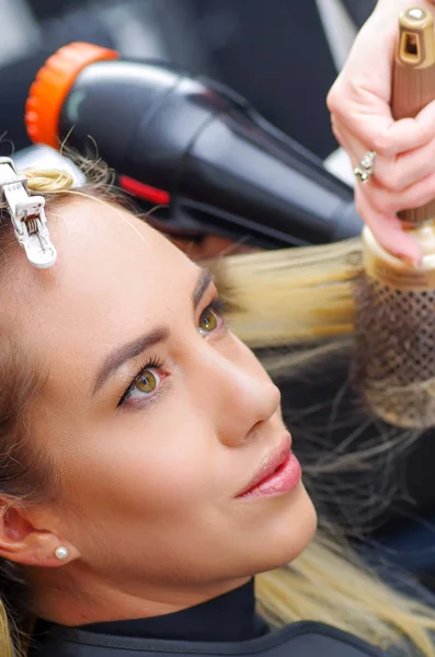 Porträtt av en vacker kvinna i en salong och hår Frisör torkning blont hår med hårtork och rund hårborste i en suddig bakgrund — Stockfoto