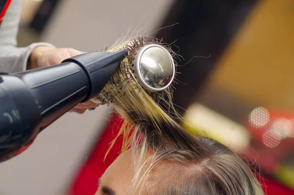 Κοντινό πλάνο της ένα στιλίστα τρίχας ξήρανση ξανθά μαλλιά με πιστολάκι και στρογγυλή βούρτσα μαλλιών σε ένα θολή φόντο — Φωτογραφία Αρχείου