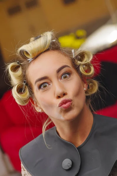 Blond žena v kadeřnictví s natáčky v hlavě, v pozadí salon vlasy, dělá legrační obličeje zblízka — Stock fotografie