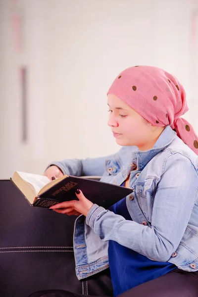 Close up de uma mulher com câncer vestindo lenço de cabeça, sentado em um sofá preto lendo um livro, em um fundo embaçado — Fotografia de Stock