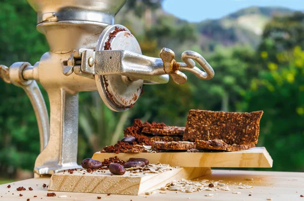 Barrette di granola con cereali e fave di cacao su un tavolo di legno e un mulino a mano grigio, in uno sfondo di natura sfocata — Foto Stock