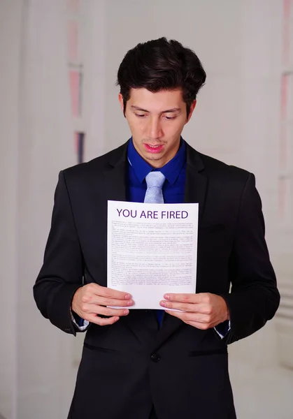 Close up of surprised businessman wearing a suit and holding a sheet of paper of youre fired text on it, in a blurred background — Stock Photo, Image