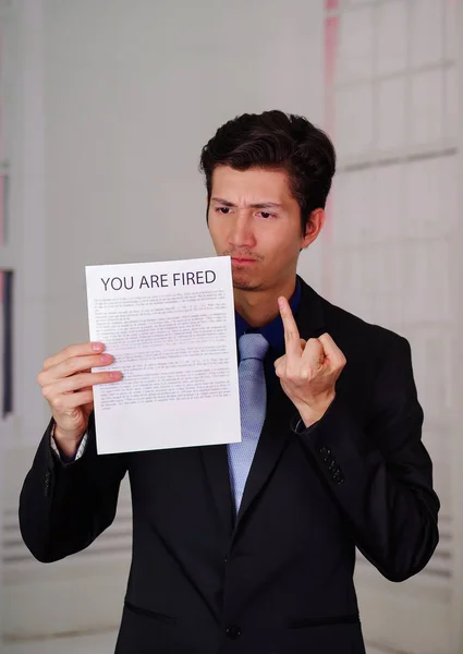 Close up of angry businessman wearing a suit and holding a sheet of paper of youre fired text on it, doing a middle finger sign, in a blurred background — Stock Photo, Image