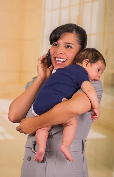 Joven madre está sosteniendo a su pequeño bebé y usando su celular hger en un fondo borroso — Foto de Stock