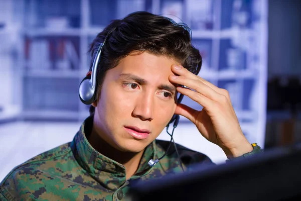 Retrato de soldado preocupado vistiendo un uniforme militar, tocando su cabeza y mirando a su computadora con sus auriculares alrededor de la hea, listo para dar un consejo, en un fondo borroso —  Fotos de Stock