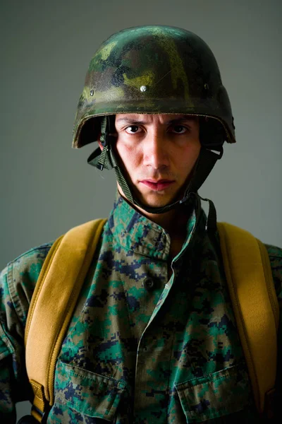 Retrato de un joven soldado serio con un uniforme militar, un casco y una mochila en un fondo gris —  Fotos de Stock