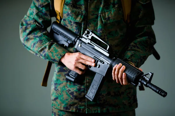 Close up de soldado segurando um rifle com as duas mãos e vestindo um uniforme militar, em um fundo cinza — Fotografia de Stock