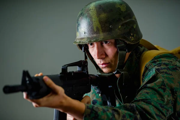 Portrait of young soldier holding in his hands a rifle, wearing a military uniform, in a gray background — Stock Photo, Image