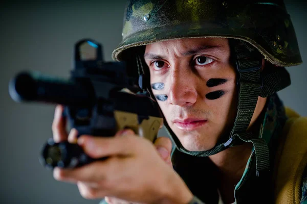 Портрет красивого молодого солдата, який тримає в руках гвинтівку, готову до стрілянини, носить військову форму, розфарбовує обличчя двома пальцями, на сірому фоні — стокове фото