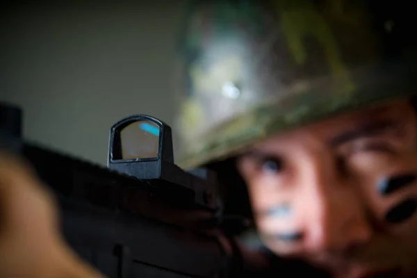 Retrato de joven soldado guapo sosteniendo en sus manos un rifle listo para disparar, vistiendo un uniforme militar, pintado su cara con dos dedos, enfoque selectivo en un fondo gris —  Fotos de Stock