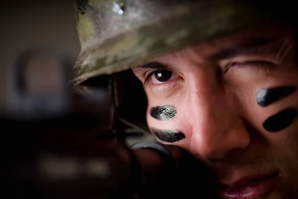 Retrato de joven soldado guapo sosteniendo en sus manos un rifle listo para disparar, guiñar el ojo para tener más precisión y usar un uniforme militar, pintado su cara con dos dedos, en un gris —  Fotos de Stock