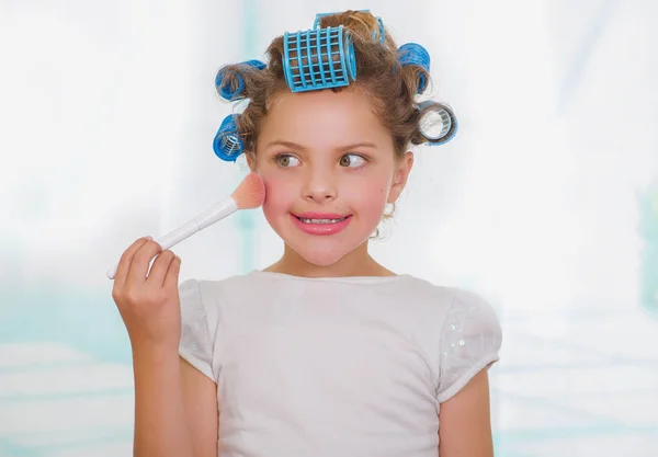 Menina usando um pincel para fazer as pazes enquanto vestindo rolos de cabelo e roupão de banho em um fundo embaçado — Fotografia de Stock