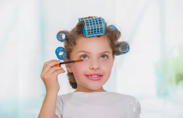 Bulanık bir arka planda saç-silindirler ve bornoz giyen süre gözüne bir göz rimel kullanan küçük kız — Stok fotoğraf