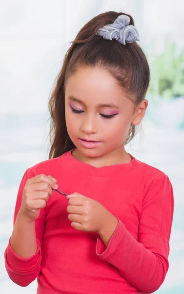 Portrait of beautiful little girl painting her nails, wearing a red blouse in a blurred background — Stock Photo, Image