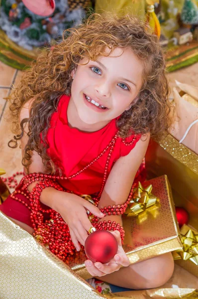 Gros plan de fille heureuse portant une robe rouge et tenant une boule de Noël et la décoration dans ses mains, avec un arbre de Noël derrière, concept de Noël — Photo