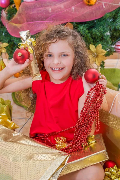 Primer plano de niña sonriente con una blusa roja y sosteniendo una bola de Navidad, decoración y regalo en sus manos, con un árbol de Navidad detrás, concepto de Navidad — Foto de Stock