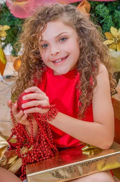 Close up van gelukkig meisje draagt een rode jurk en holding een kerst bal en decoratie in haar handen, met een kerstboom achter, Kerstmis concept — Stockfoto