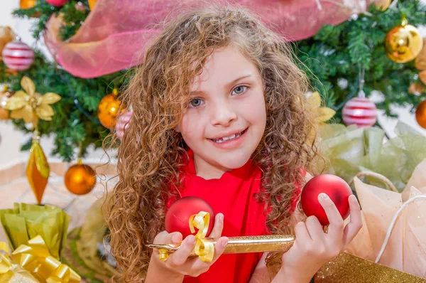 Close up van lachende meisje draagt een rode blouse en holding een kerst bal, decoratie en cadeau in haar handen, met een kerstboom achter, Kerstmis concept — Stockfoto