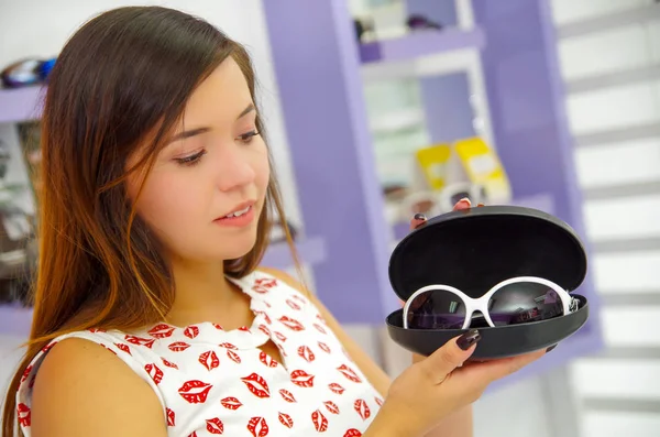 Belle jeune femme tenant dans ses mains des lunettes de soleil en magasin d'optique, en arrière-plan flou — Photo