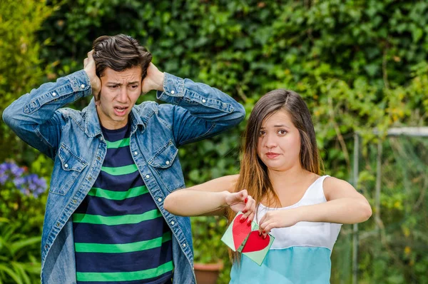 Close up van een jonge vrouw met een afschuwelijk gezicht en scheuren van een brief die zij houdt in haar handen, met een bezorgd vriendje met beide handen in zijn hoofd die op zoek zijn vriendin, vriend zone concept — Stockfoto