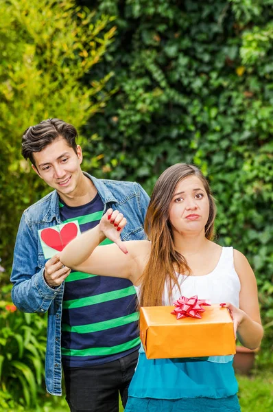 Close up of young woman doing a face of detesting the gift she is holding in her hands and doing a thumbs down, with smiling boyfriend behind holding a love letter looking his girlfriend, friend zone — Stock Photo, Image