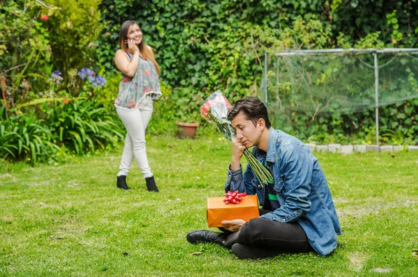 Close up of sad man wearing a jean jacket and black pants sitting in the ground holding gift with a blurred woman behind using her cellphone. Valentines Day concept or friend zone concept