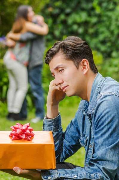 Primer plano de un hombre triste con una chaqueta de jean y pantalones negros sentados en el suelo sosteniendo un regalo en sus manos, con una pareja borrosa detrás abrazándose. Concepto de San Valentín o zona de amigos —  Fotos de Stock