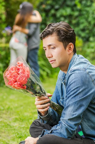 Gros plan d'un homme triste portant une veste en jean et un pantalon noir assis dans le sol tenant des fleurs dans ses mains, avec un couple flou derrière s'embrasser. Concept de Saint-Valentin ou zone ami — Photo