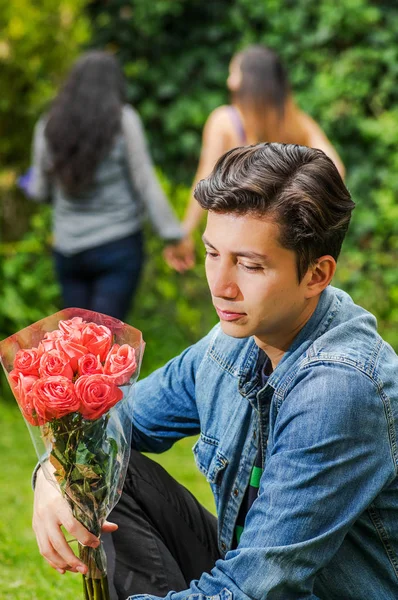 Primer plano de un hombre triste con una chaqueta de jean y pantalones negros sentados en el suelo sosteniendo flores en sus manos, con una pareja lesbiana borrosa detrás sosteniendo sus manos. Concepto de San Valentín o —  Fotos de Stock