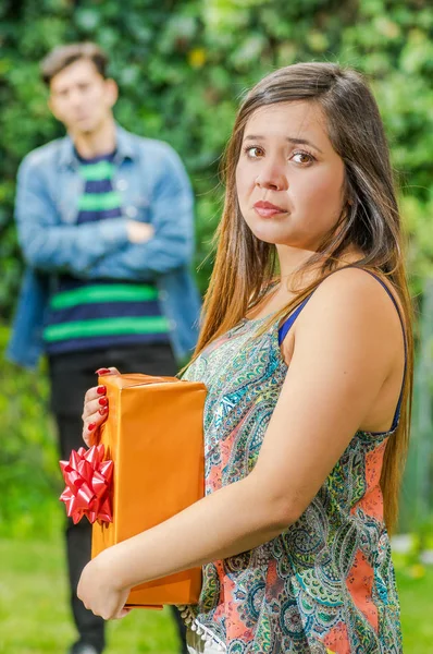 Close up of sad woman holding a gift in her hands, with her endless love man behind. Valentines Day concept or friend zone concept