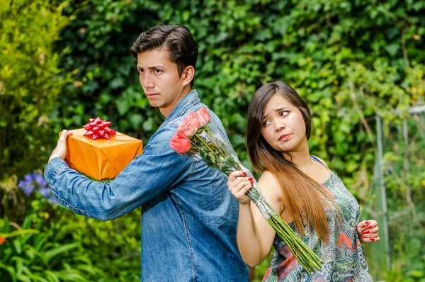 Close up of mad woman holding flowers and mad man holding a gift back to back ignoring each other, friend zone concept — Stock Photo, Image