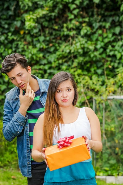 Nahaufnahme einer jungen Frau, die das Geschenk, das sie in den Händen hält, verabscheut, mit einem nachdenklichen Freund im Rücken und besorgtem Gesicht, Freund-Zone-Konzept — Stockfoto